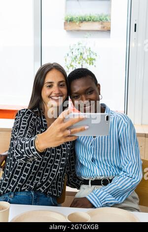 Des collègues féminins gais et multiraciaux se font autoportrait sur leur smartphone tout en déjeunant ensemble dans leur cuisine sur leur lieu de travail Banque D'Images