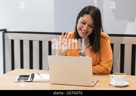 Une femme pigiste ravie de se faire passer la main et de passer des appels vidéo sur netbook tout en travaillant à distance dans un café Banque D'Images