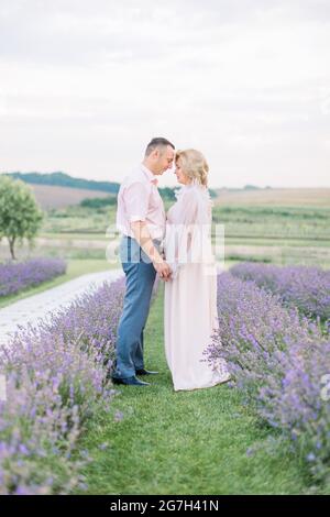 Portrait de famille d'un homme et d'une jolie femme d'âge moyen, debout à l'extérieur sur un champ de lavande, tenant les mains et touchant les fronts. Concept anniversaire de mariage. L'amour à travers les années Banque D'Images