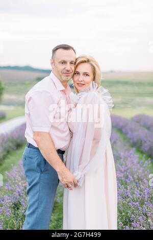 Vue rapprochée de l'avant du charmant couple mature au champ de lavande, en tenant les mains et en s'embrassant. Photo de l'homme et de la femme qui s'occupent de marche à l'extérieur dans le champ de lavande Banque D'Images