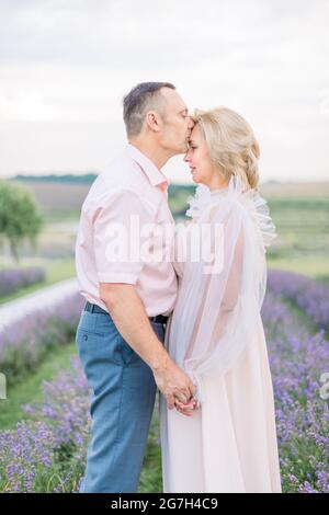 Couple amoureux mûr et romantique marchant dans le champ de lavande. Couple d'âge moyen de race blanche rêveuse et heureuse dans la nature, debout tenant les mains. Homme embrassant sa femme. Profitez de l'instant présent. Banque D'Images