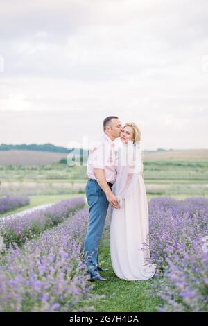 Couple amoureux mûr et romantique marchant dans le champ de lavande. Couple d'âge moyen de race blanche rêveuse et heureuse dans la nature, debout tenant les mains. Homme embrassant sa femme. Profitez de l'instant présent. Banque D'Images