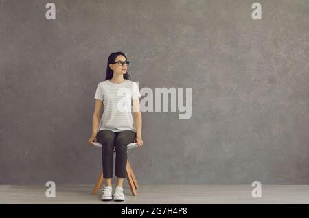 Une jeune fille belle est assise sur un tabouret blanc en pleine croissance, dans un T-shirt, chaussures de sport. Banque D'Images