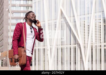 homme d'affaires noir souriant avec porte-documents traverse le centre-ville financier en parlant au téléphone, concept de technologie et de communication, copy spa Banque D'Images