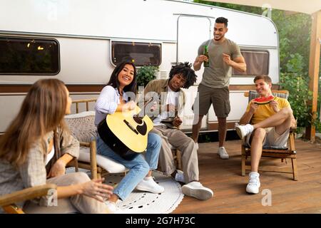 Groupe de jeunes amis divers ayant une fête en plein air près de RV, jouant de la guitare, chantant, boire de la bière, s'amuser ensemble. Millénn multiracial joyeux Banque D'Images