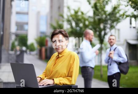 Concept de l'industrie des affaires et de la finance. Jeune femme d'affaires attirante dans un chemisier jaune debout à l'extérieur dans le centre-ville à l'aide d'un ordinateur portable et regardant directement à l'appareil photo. Deux hommes parlent en arrière-plan. Banque D'Images