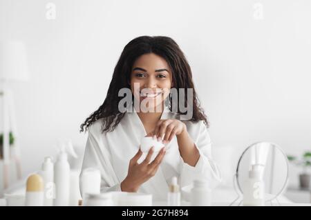 Routine du matin. Belle femme afro-américaine en peignoir après avoir appliqué la crème hydratante sur le visage, tenant le pot avec la lotion, assis dans Banque D'Images