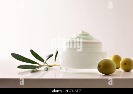 Crème cosmétique hydratante à l'extrait d'olive pour la peau avec branche et olives sur table blanche. Vue avant. Banque D'Images