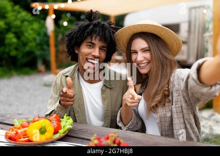 Jeune couple multinational déjeuner ensemble au camping, posant pour selfie près de la camionnette de camping, pointant vers l'appareil photo et souriant. Cool Black Guy et salut Banque D'Images