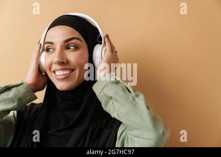 Un portrait de la femme musulmane souriante portant le hijab noir et tenant les écouteurs tout en se tenant dans le studio orange Banque D'Images