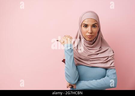 Une femme musulmane portant le hijab rose pointant vers le copyspace dans le studio rose Banque D'Images