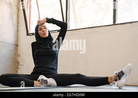 Jeune sportif musulman dans le hijab se reposant tout en faisant de l'activité à l'intérieur Banque D'Images