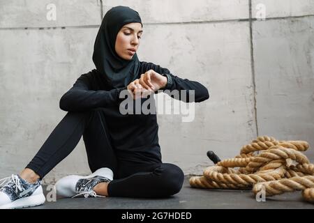 Jeune femme musulmane dans le hijab regardant la montre intelligente tout en étant assise sur le sol à l'intérieur Banque D'Images