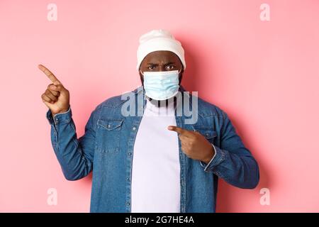 Coronavirus, mode de vie et concept de pandémie mondiale. Un homme afro-américain en colère et déçu, dans un masque facial pointant vers la gauche, regardant la caméra Banque D'Images
