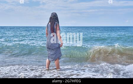Une jeune femme avec des dreadlocks sur les cheveux noirs, en casquette de baseball et longue robe rayée marchant dans l'eau. Banque D'Images