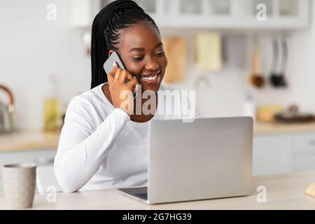 Activités à distance. Portrait de la jeune femme africaine en congé de maternité parlant sur téléphone portable assis à la table dans la cuisine avec ordinateur portable, travaillant à distance Mak Banque D'Images