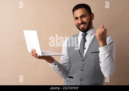 Joyeux jeune homme arabe beau travaillant sur un ordinateur portable tout en se tenant sur un fond beige Banque D'Images