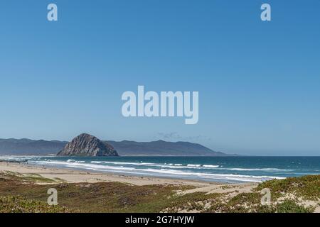 Morro Bay, CA, Etats-Unis - juin 10 2021 : Morro Rock gris foncé dans l'océan Pacifique bleu profond sous le ciel bleu avec chaîne de montagnes à l'horizon. Dunes couvertes de vert Banque D'Images