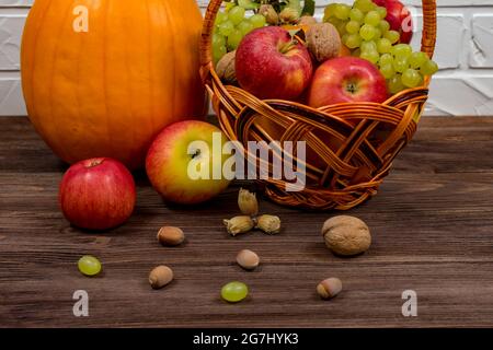 Citrouilles orange avec pommes et poires dans un panier en osier avec raisins, noix et feuilles sur une table en bois marron sur fond de mur en brique blanche Banque D'Images