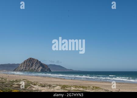 Morro Bay, CA, Etats-Unis - juin 10 2021 : Morro Rock gris foncé dans l'océan Pacifique bleu profond sous ciel bleu. Dunes couvertes de vert et sable à l'avant. Surf blanc. Banque D'Images