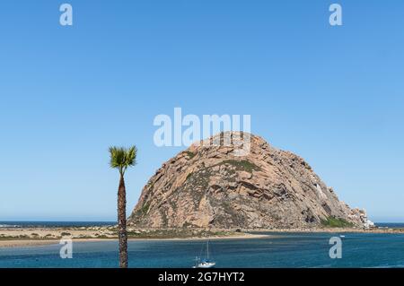 Morro Bay, CA, Etats-Unis - juin 10 2021 : Morro Rock brun dans l'océan Pacifique bleu profond sous le ciel bleu vu du centre-ville avec un bateau sur la marina et le palmier. Sa Banque D'Images