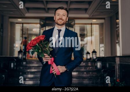 Habillé de manière formelle, homme d'affaires attrayant attendant une petite amie avec bouquet de roses Banque D'Images