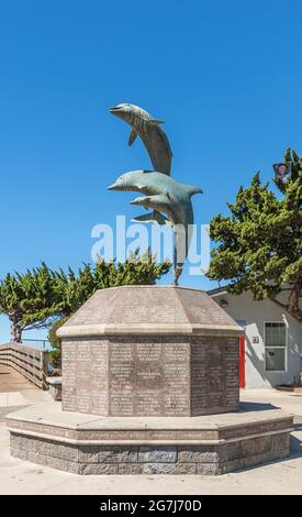 Cayucos, CA, USA - 10 juin 2021 : statue de dauphins sautants sur un piédestal en pierre brune avec banc circulaire à la base de la jetée contre le ciel bleu. Feuillage vert Banque D'Images