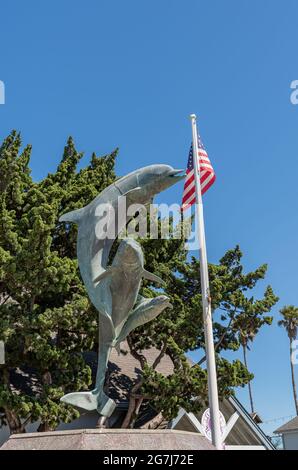 Cayucos, CA, Etats-Unis - 10 juin 2021 : gros plan des dauphins sautants sur la statue et le drapeau américain et le feuillage vert sous le ciel bleu. Banque D'Images