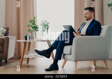 Homme d'affaires réussi en tenue habillée assis dans un fauteuil confortable utilise un ordinateur portable moderne prépare des poses de projet en ligne contre les analyses de l'intérieur de la maison Banque D'Images