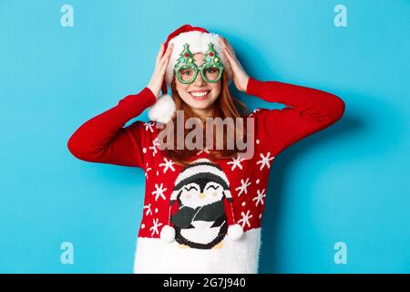 Joyeuses fêtes et concept de Noël. Drôle redhead adolescente célébrant le nouvel an, portant chapeau de santa et lunettes de fête, debout contre le bleu Banque D'Images