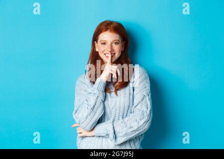 Un adolescent assez rouge se précipitant et souriant, racontant un secret, debout dans un chandail sur fond bleu Banque D'Images