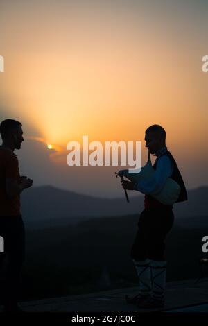 Un jeune joueur de cornemuse en costume folklorique joue sur un pic de montagne quand le soleil se lève, le matin 01 juillet 2021, Shipka, Bulgarie Banque D'Images