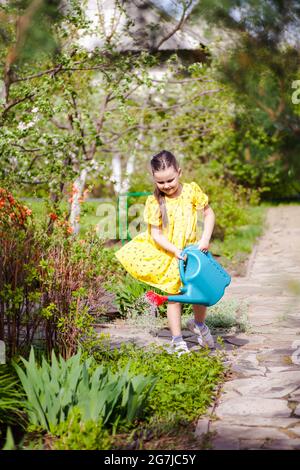 une fille souriante, joyeuse dans une robe jaune volant dans le vent est arroser de jeunes fleurs d'un arrosoir bleu dans la cour avant d'une maison dans un Banque D'Images