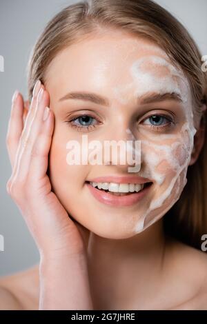jeune femme heureuse avec des yeux bleus et de la mousse nettoyante sur le visage isolé sur le gris Banque D'Images