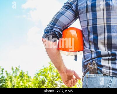 Homme attrayant dans des vêtements de travail, tenant des outils dans ses mains contre le fond des arbres, le ciel bleu et le coucher du soleil. Vue de l'arrière. Main-d'œuvre et emploi Banque D'Images