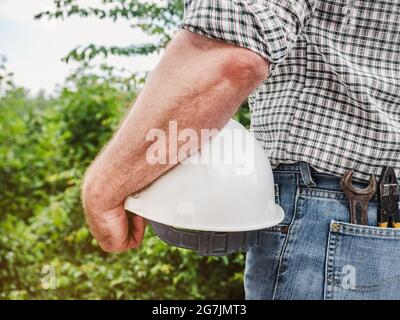 Homme attrayant dans des vêtements de travail, tenant des outils dans ses mains contre le fond des arbres, le ciel bleu et le coucher du soleil. Vue de l'arrière. Main-d'œuvre et emploi Banque D'Images