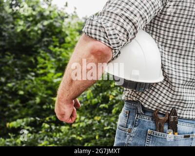 Homme attrayant dans des vêtements de travail, tenant des outils dans ses mains contre le fond des arbres, le ciel bleu et le coucher du soleil. Vue de l'arrière. Main-d'œuvre et emploi Banque D'Images