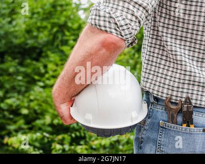 Homme attrayant dans des vêtements de travail, tenant des outils dans ses mains contre le fond des arbres, le ciel bleu et le coucher du soleil. Vue de l'arrière. Main-d'œuvre et emploi Banque D'Images