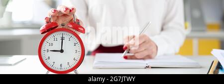 Une femme d'affaires à son bureau tient la main sur l'alarme rouge horloge et effectue des entrées dans le journal Banque D'Images