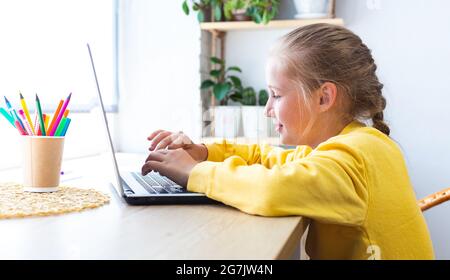 Blanche blonde école fille, tapant sur un clavier d'ordinateur portable tout en étant assis près de la fenêtre à la maison, blogging, chat, affichage des pages de médias sociaux ou Banque D'Images