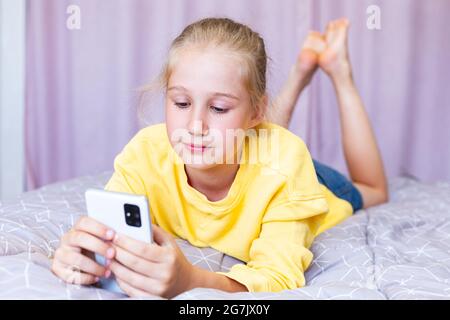 Une jeune fille blanche blonde se trouve sur le lit avec un téléphone dans ses mains, joue à des jeux, magasine en ligne, blogging. Communiquer sur les réseaux sociaux Banque D'Images