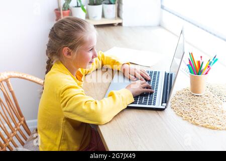 Blanche blonde école fille, tapant sur un clavier d'ordinateur portable tout en étant assis près de la fenêtre à la maison, faire des devoirs, blogging, chat, regarder social Banque D'Images