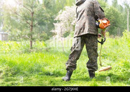 Un homme qui travaille en camo porte la tonte de l'herbe haute avec un coupe-herbe à essence ou une tondeuse à gazon dans le parc de la ville, le jardin ou la cour. Outils d'entretien de jardinage et Banque D'Images
