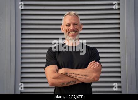 Portrait d'un homme mûr en forme parfaite souriant à l'appareil photo tout en se tenant debout avec les bras croisés à l'extérieur sur fond gris Banque D'Images