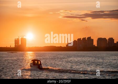 Silhouettes de bâtiments de la ville sur la rive contre le fond du soleil couchant et de l'éblouissement sur l'eau. Coucher de soleil sur la rivière, un bateau à moteur traverse Banque D'Images