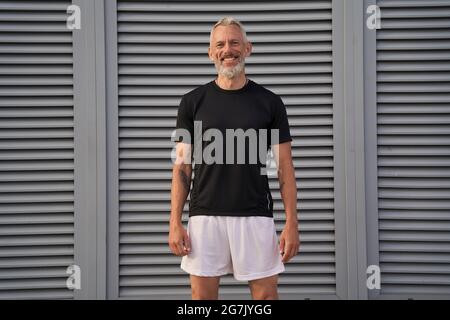Heureux homme d'âge moyen en forme parfaite souriant à l'appareil photo tout en se tenant à l'extérieur après l'entraînement Banque D'Images
