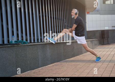 Prise de vue en longueur d'un homme sportif d'âge moyen, avec des vêtements de sport et des écouteurs qui s'éloignent, réchauffent son corps et se prépare à l'entraînement en plein air Banque D'Images