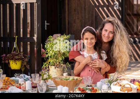 Portrait d'une belle femme souriante embrassant sa petite fille de derrière lorsqu'ils sont debout à la table du dîner Banque D'Images