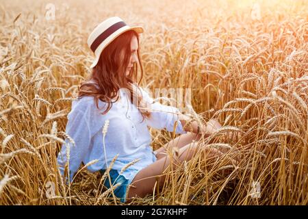 Happy Girl est assis dans un champ de blé en chapeau de paille. Mignon jeune femme d'origine caucasienne en vêtements décontractés aime le blé doré mûr dans un champ. Concept de femme libre Banque D'Images