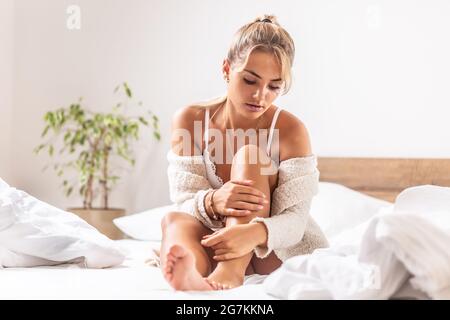 La jeune fille blonde à la peau douce applique un hydratant sur ses jambes alors qu'elle est assise sur son lit entouré de draps blancs. Banque D'Images